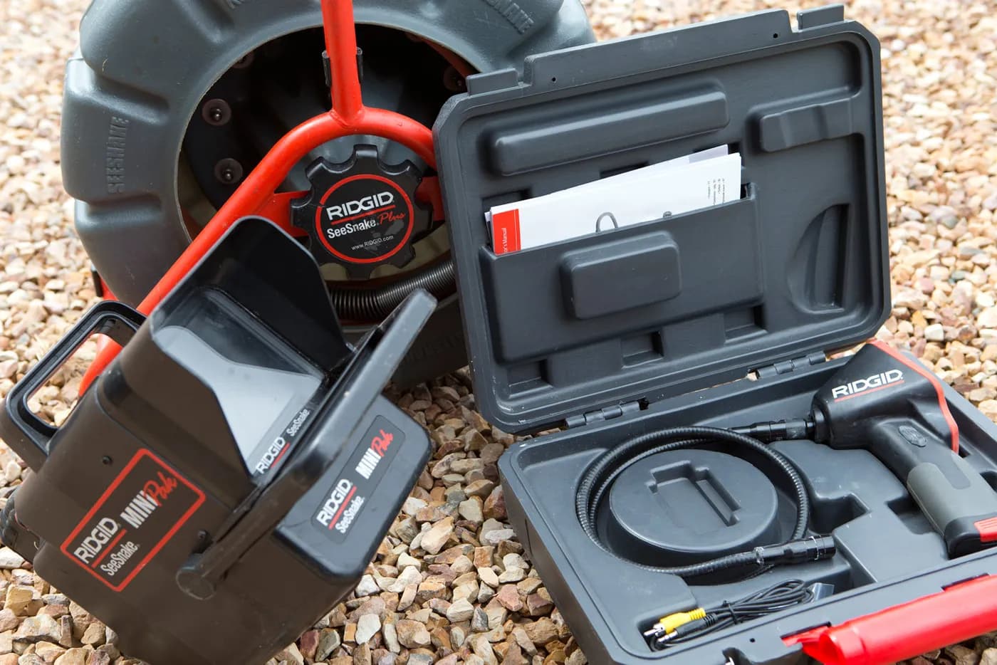 Two drain inspection camera reels and a handheld monitor on gravel, beside an open case holding a camera head and its lead