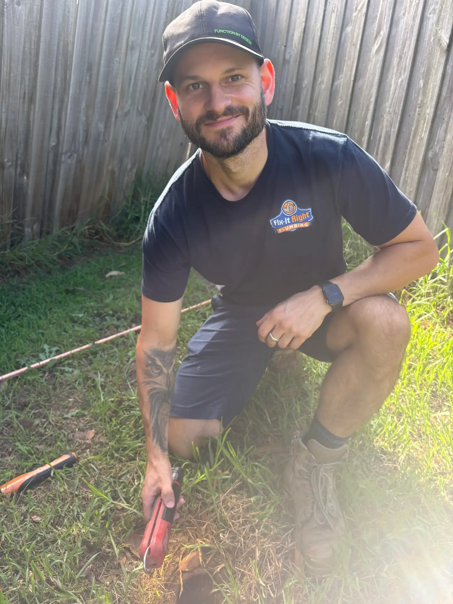 Jarryd kneeling in the grass by a timber fence with a red tool in his hand, working on the ground
