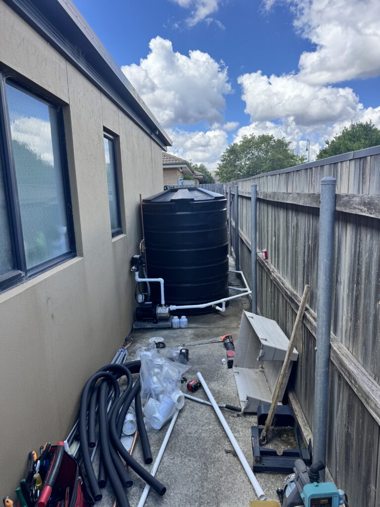 A black rainwater tank with connected piping sits on a concrete path beside a house, surrounded by various plumbing tools and materials—ideal for doomsday preppin’ or adding some stress relief through self-sufficiency.