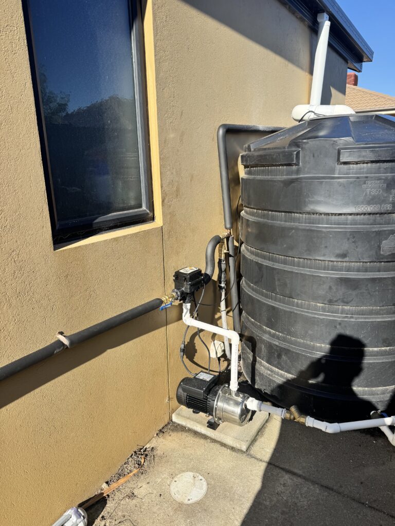 A black water tank with attached pump and pipes stands next to a tan building wall with a window, supporting emergency preparedness. A person's shadow is visible on the ground.