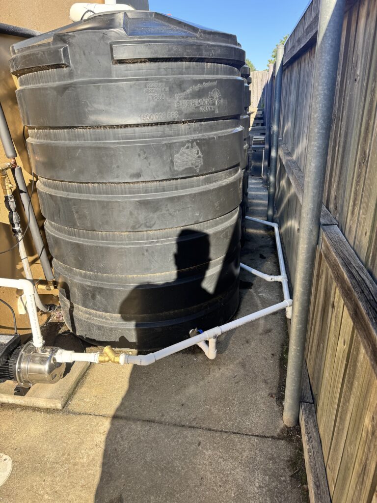Large black water tank connected to white PVC pipes and a pump, positioned between a building and a wooden fence on concrete—ideal for doomsday preppin or stress relief through reliable water storage.