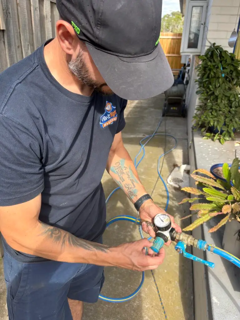 A man wearing a cap and blue shirt uses a pressure gauge to check a water pipe outside near a house and garden, part of the Bellamy Package inspection.
