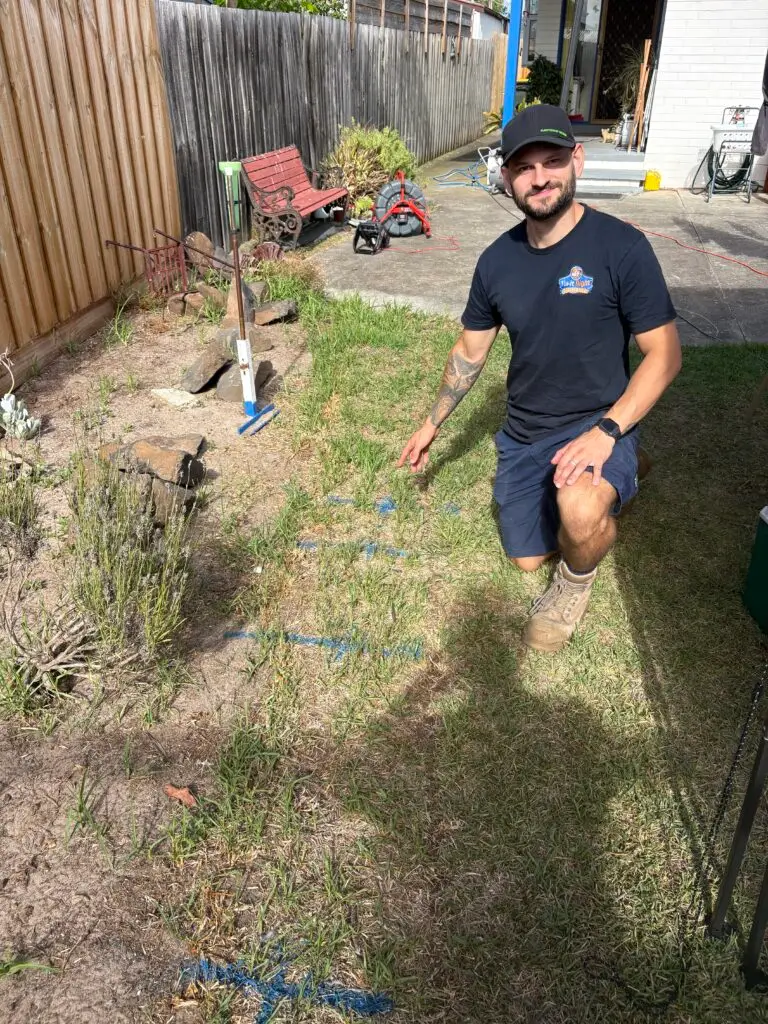 A man in work clothes kneels on grass next to a garden bed, pointing at blue spray-painted lines on the ground—part of the Craig Bellamy Package. Tools and outdoor items are visible in the background.