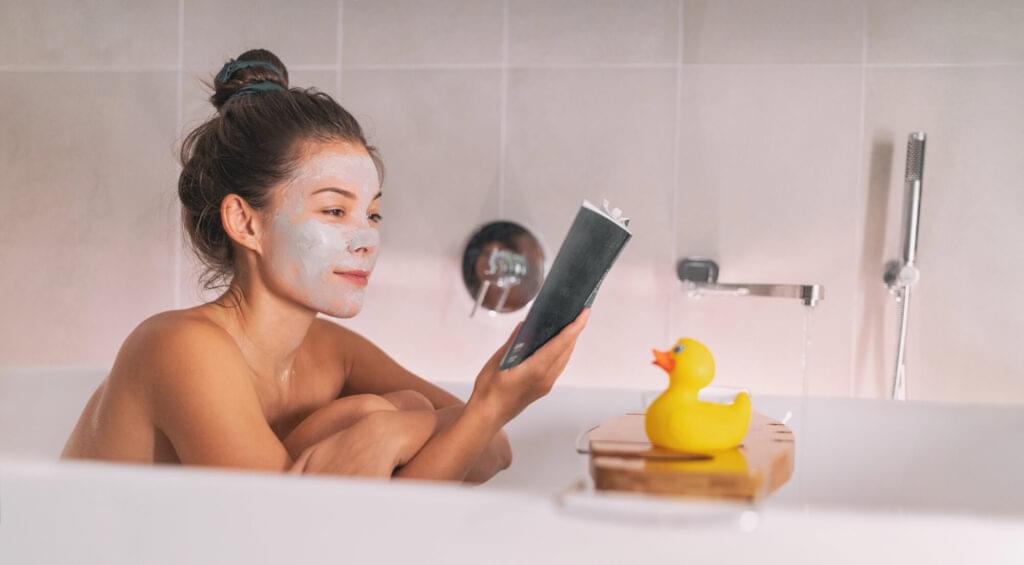 girl running a bath at home applying clay mask and reading a book soaking in hot water with rubber duck toy on bathtub caddy